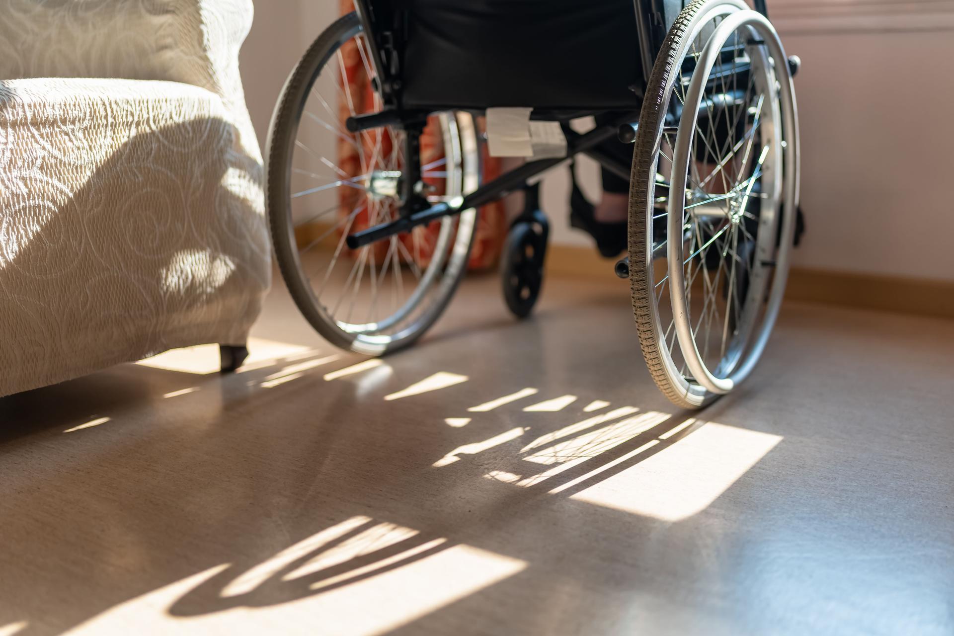 Detail of a wheelchair in front of a window and an elderly woman sitting, while enjoying the sun's rays.